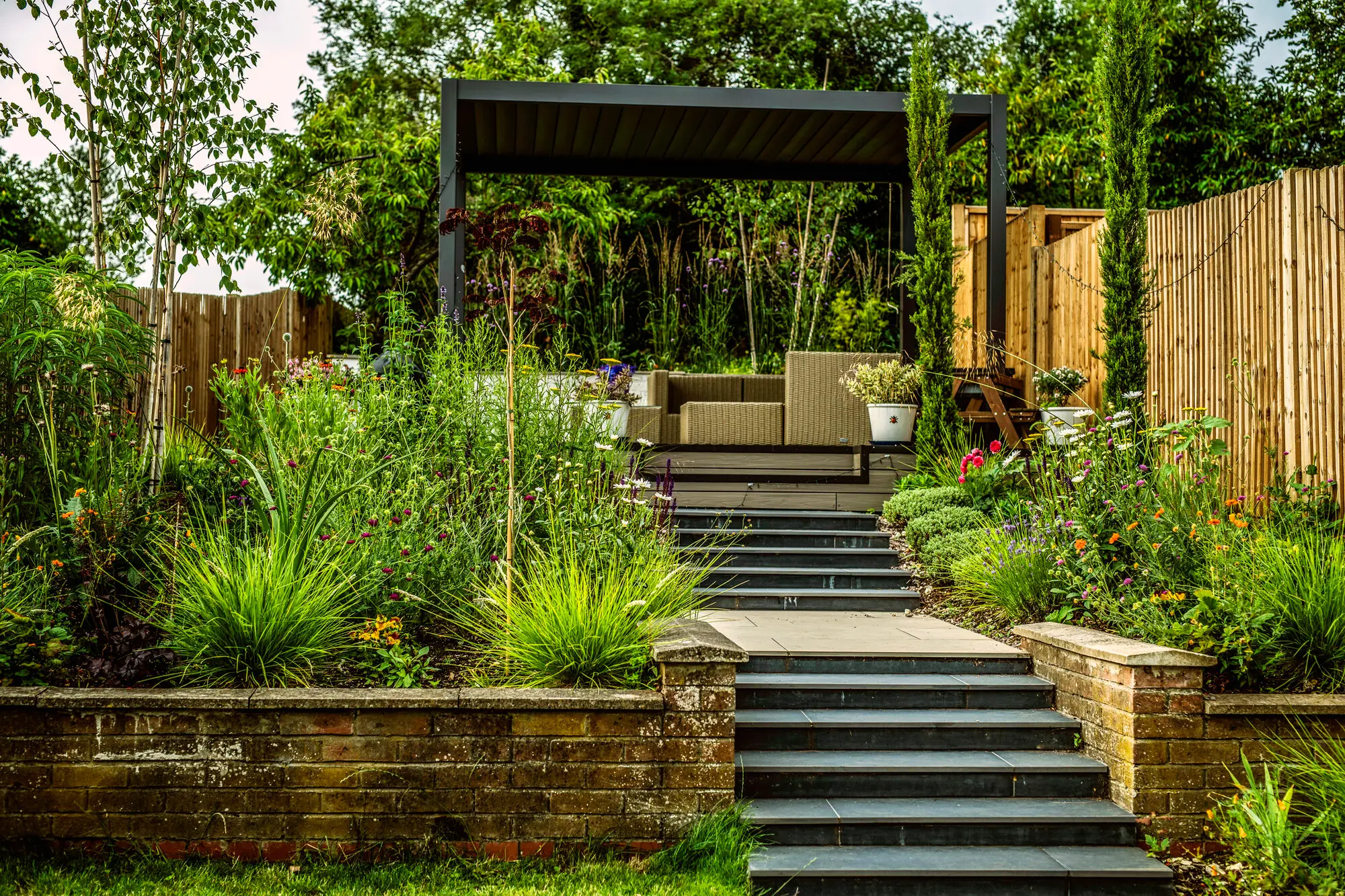 Dark stone steps rising between planted terraces to the pergola and the seating under it