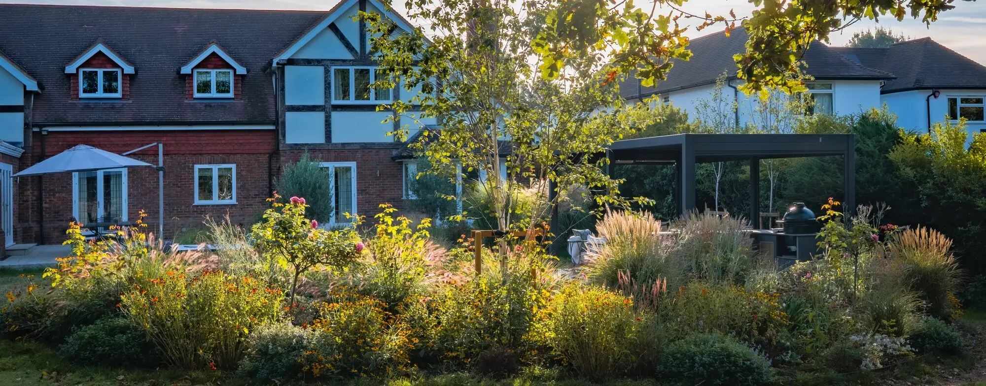 Deep borders of rudbeckia, grasses and roses in low evening sun in front of a timber-framed house, with a dark pergola and outdoor kitchen beside it