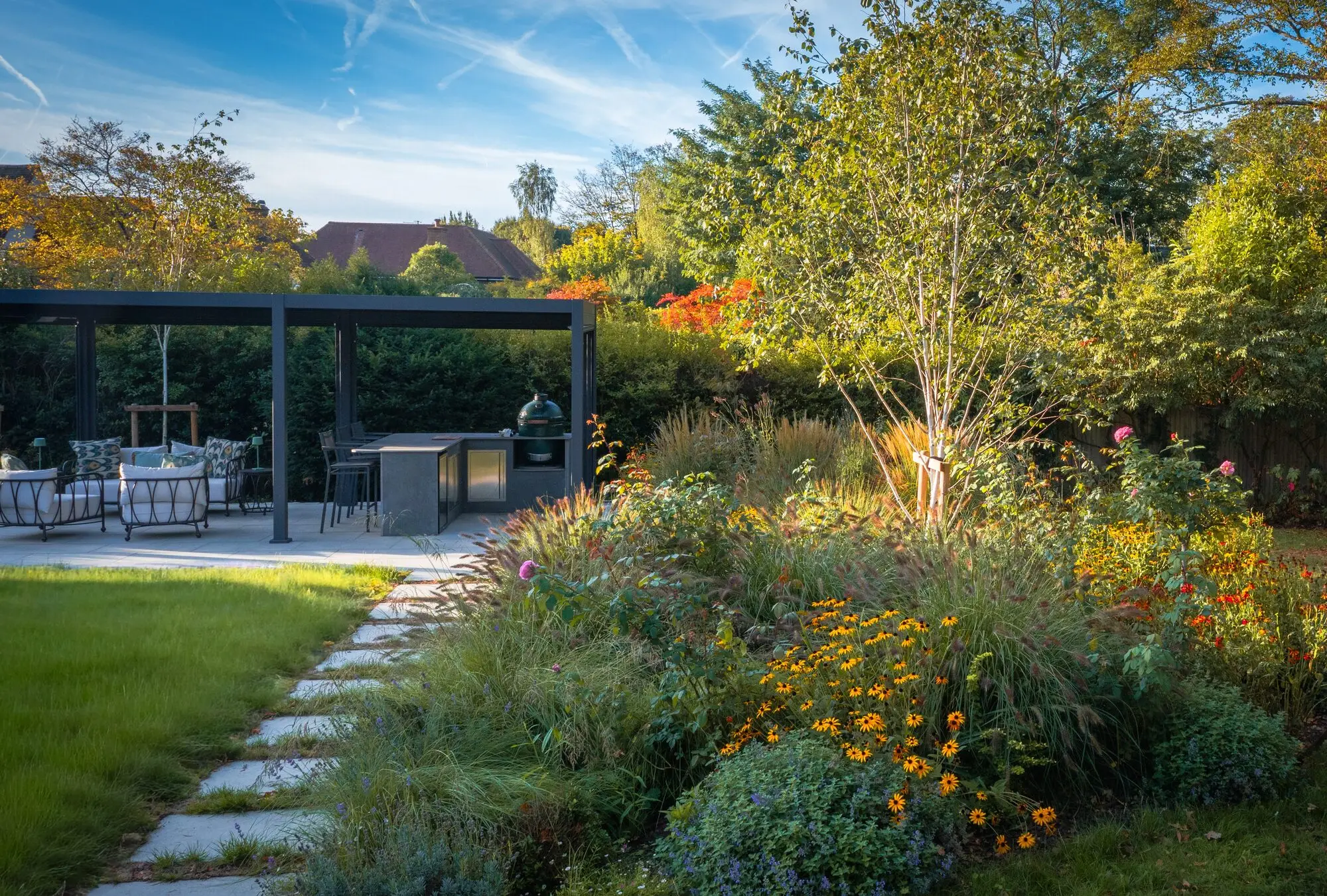 Stepping stones along the lawn beside a border of roses, rudbeckia and grasses, with the pergola behind