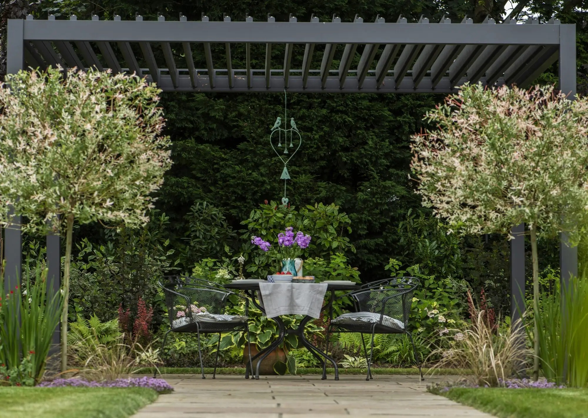 A bistro table laid for two under the grey louvred pergola, between two standard harlequin willows, with purple stocks in a vase and a heart wind chime hanging above