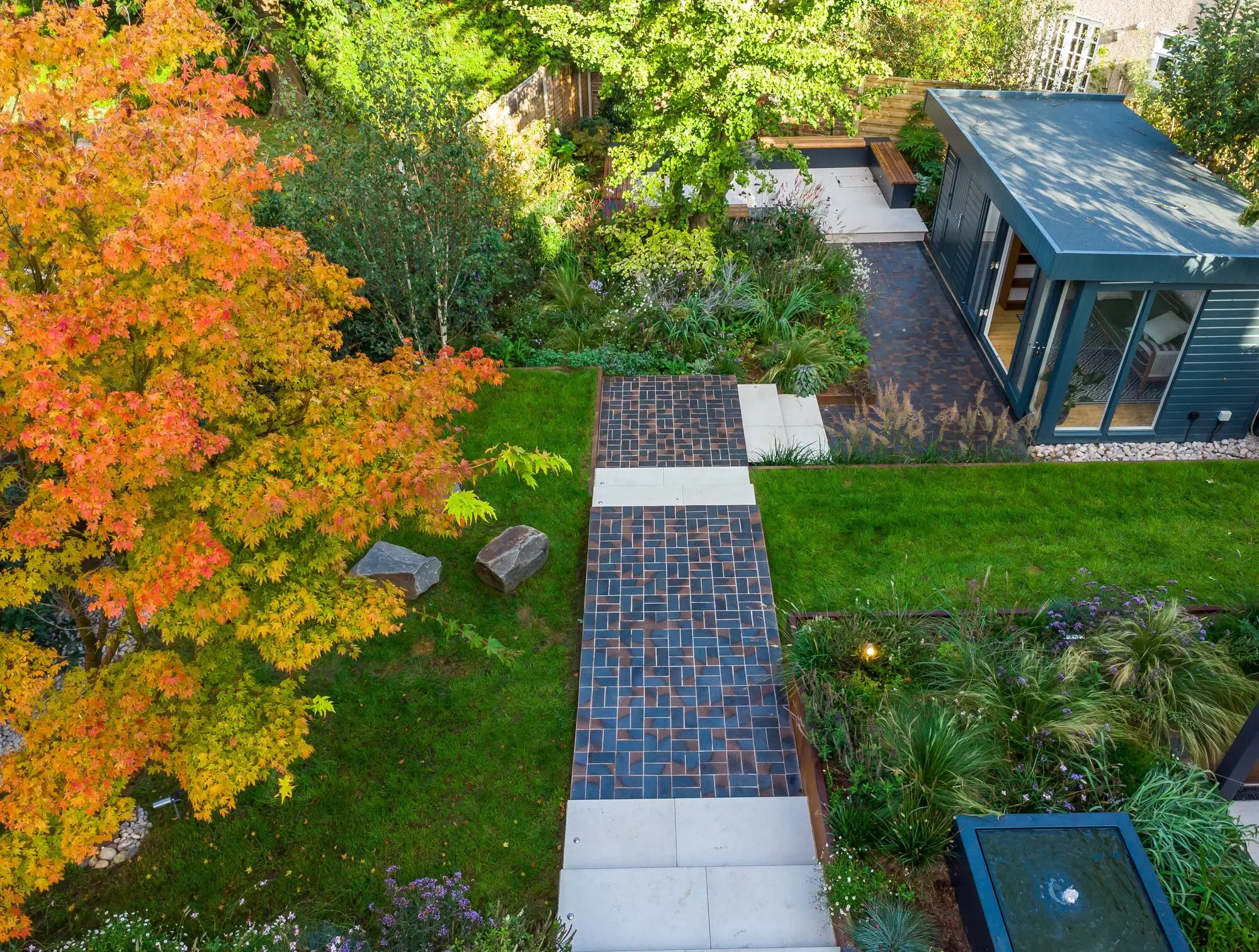 The whole garden from above, showing the lawn, the brick path, the planted terraces and the paved terrace beside the garden room, with an acer in autumn colour