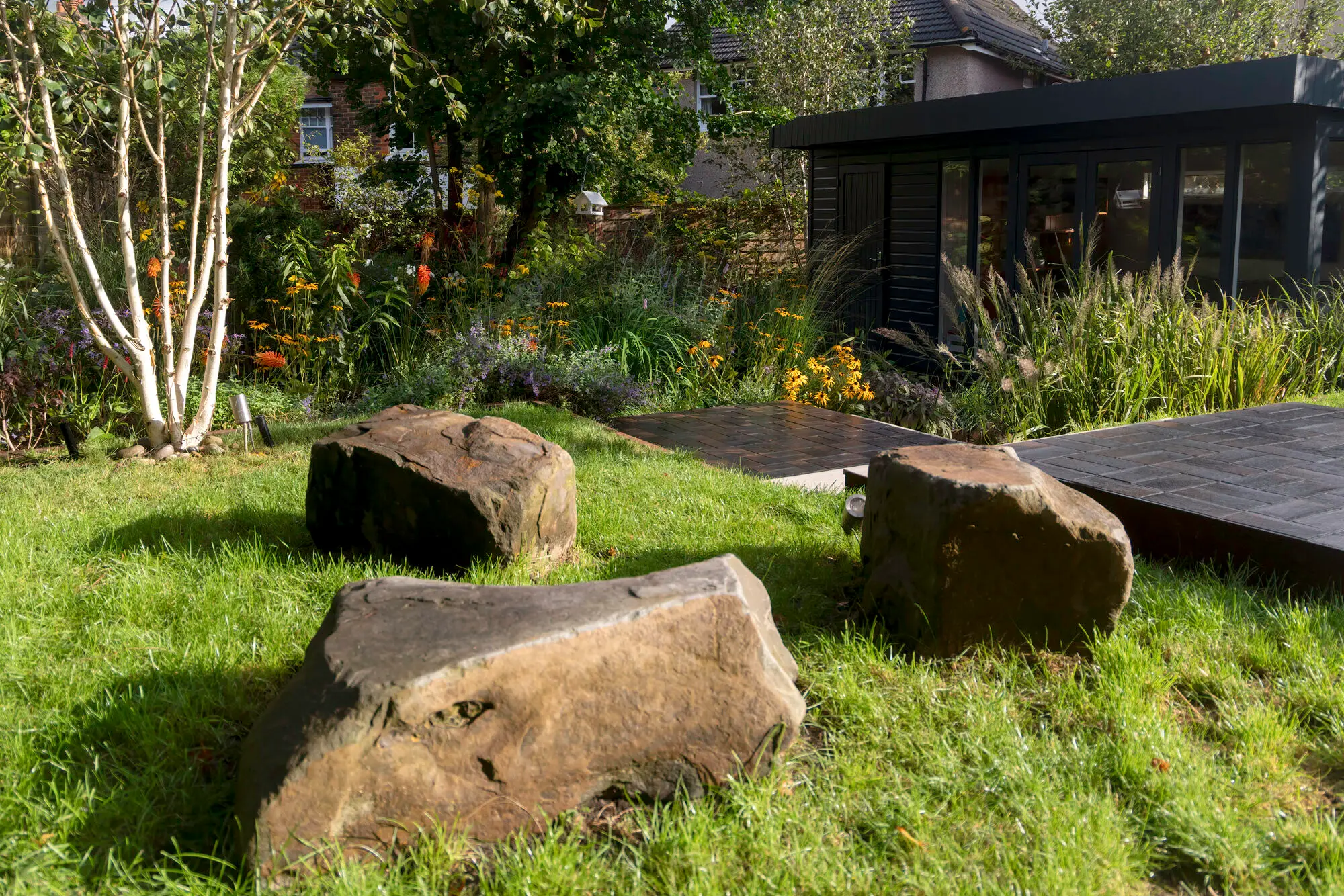 Three boulders set into the lawn beside a white-stemmed birch, with the dark garden room and its planting behind