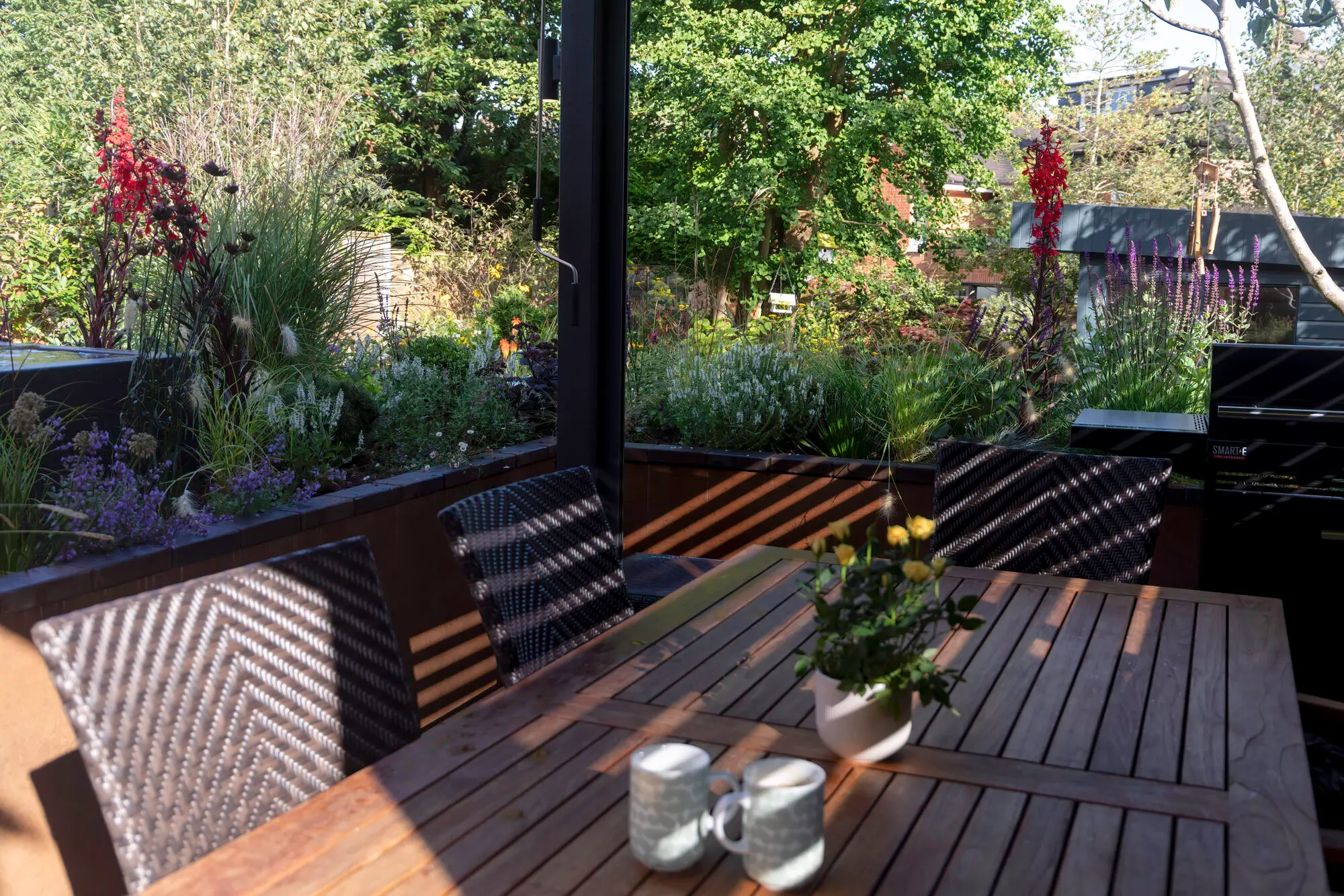 The dining table under the pergola in low afternoon sun, looking out over the perennials to the trees beyond