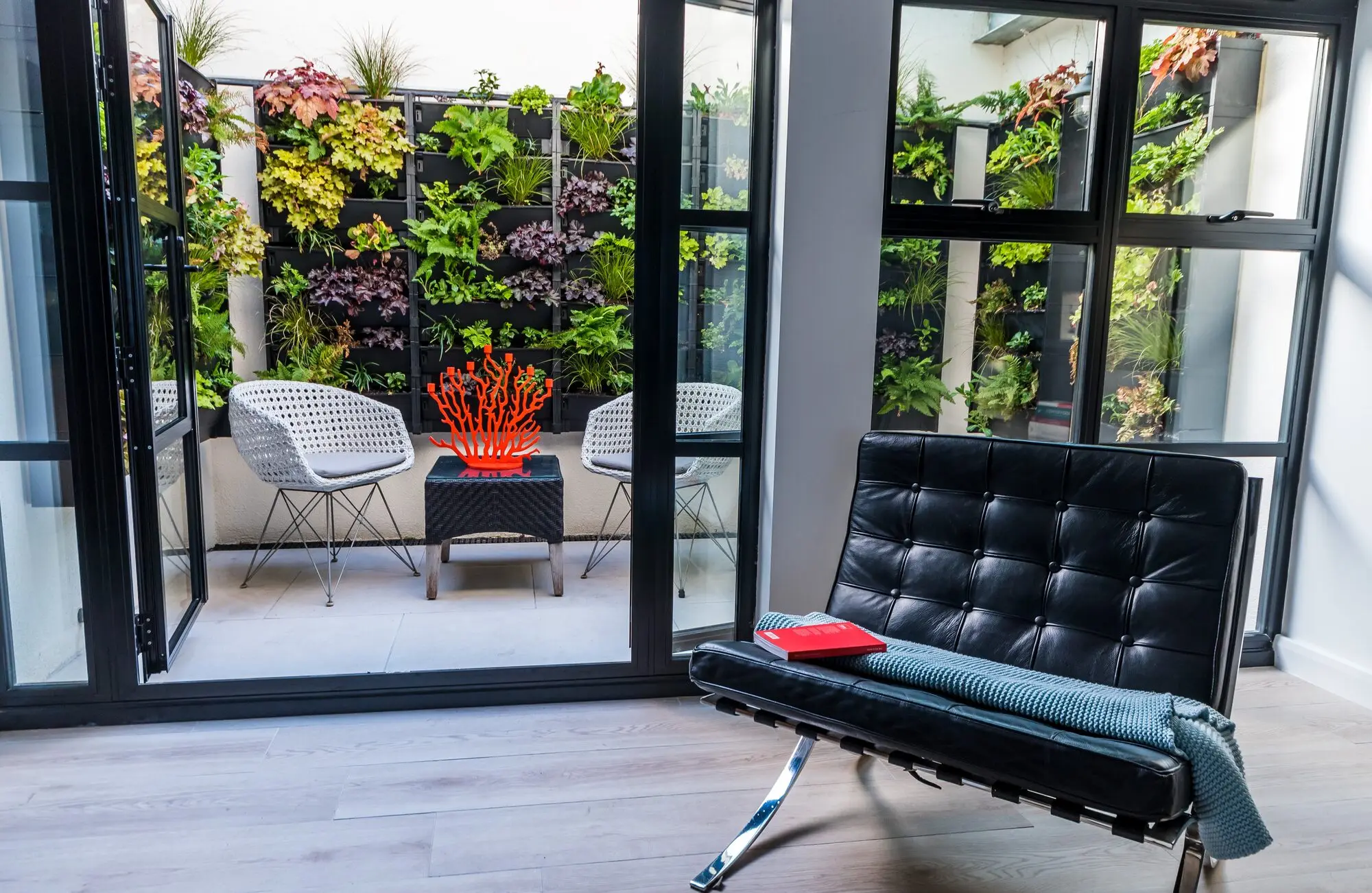 A light well planted as a living wall of ferns, heucheras and grasses, seen through steel-framed glass doors from a room with a black leather chair and a red book