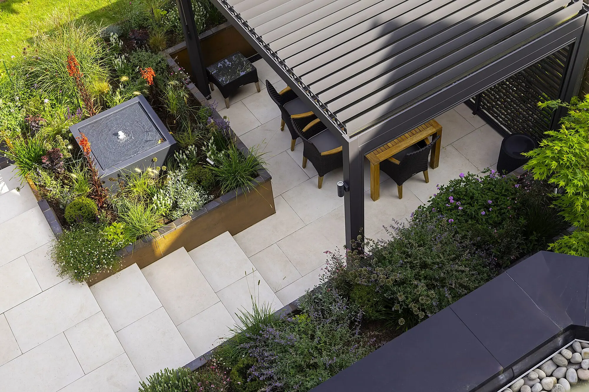 The sunken terrace from above: dining table and chairs under the louvred pergola, pale porcelain paving and steps, and a corten bed of red hot pokers around a dark square water feature