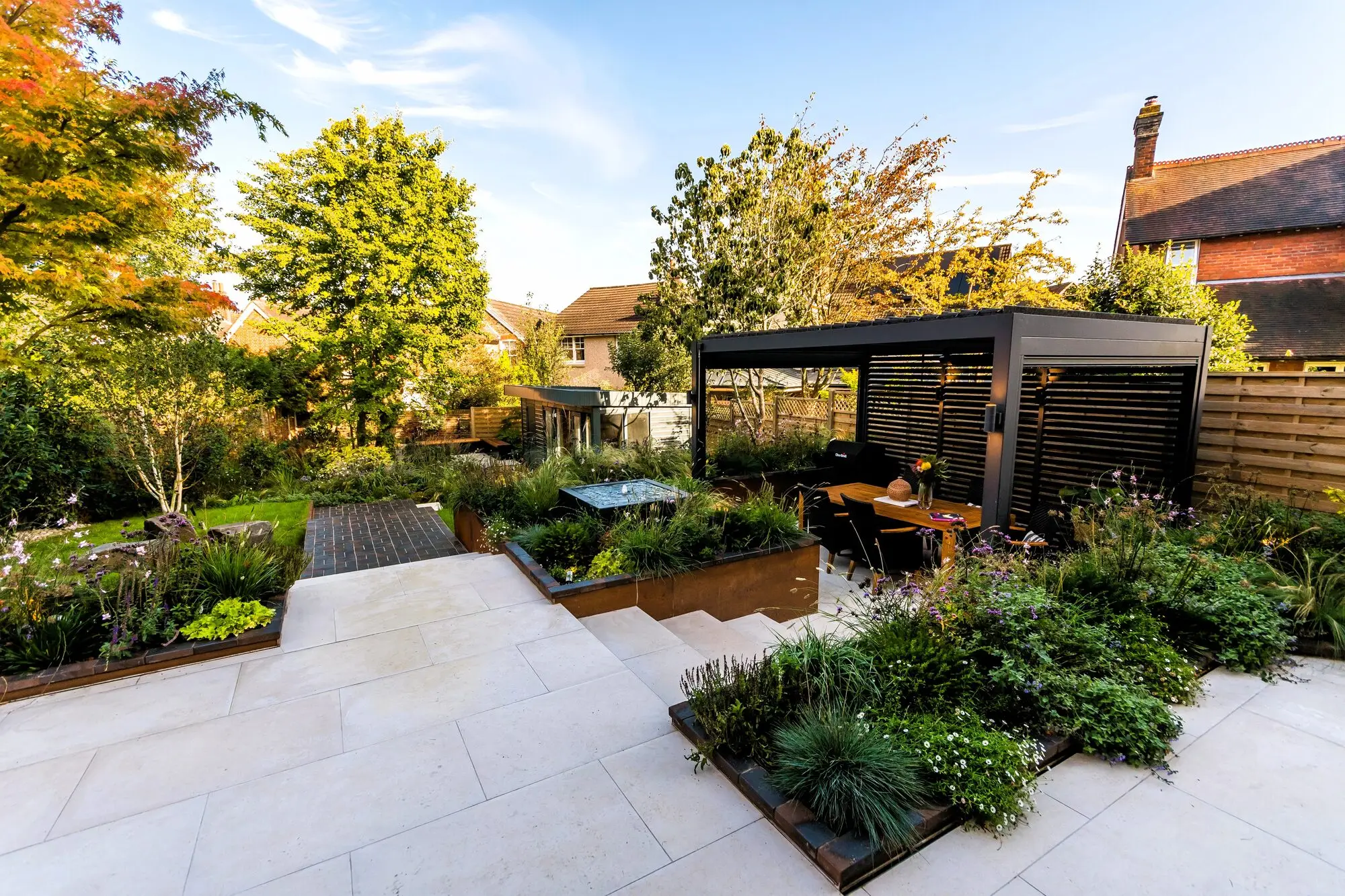 The paved upper terrace with its slatted pergola, dining table and corten-edged beds, and the aluminium water feature in a raised bed