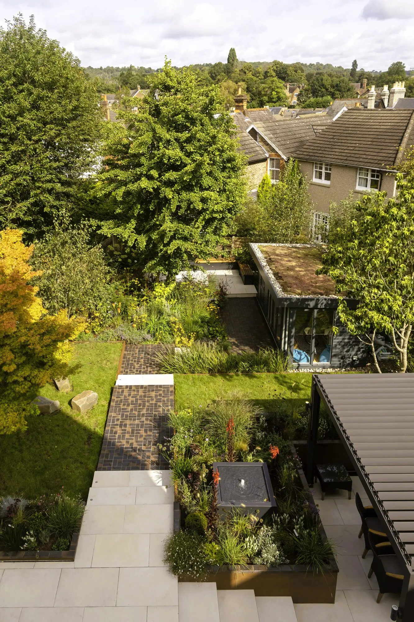 The garden in summer from the top of the house: the porcelain terrace and pergola, a clay paver path across the lawn, and the garden room with its sedum roof at the far end under the trees