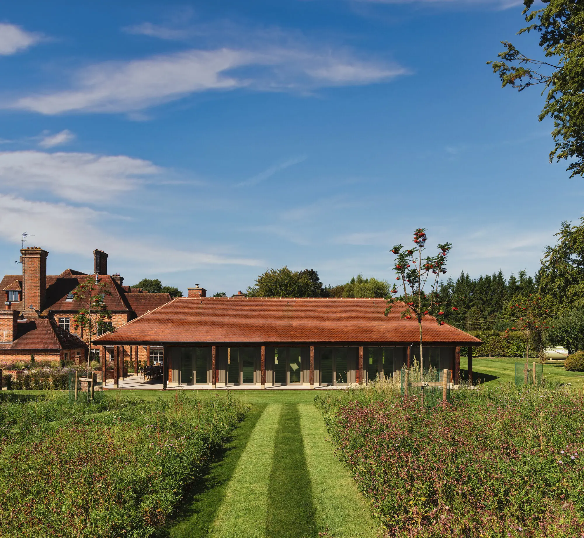 The mown lawn path running between wildflower beds to the long tiled-roof loggia, with the brick house beyond it
