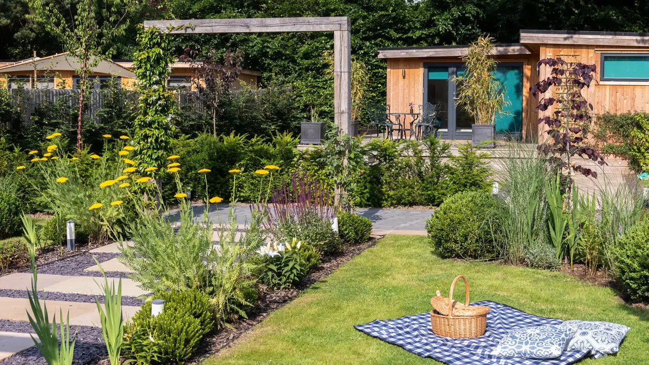 The garden room at the end of the lawn, its doors painted turquoise, with a picnic blanket laid out on the grass