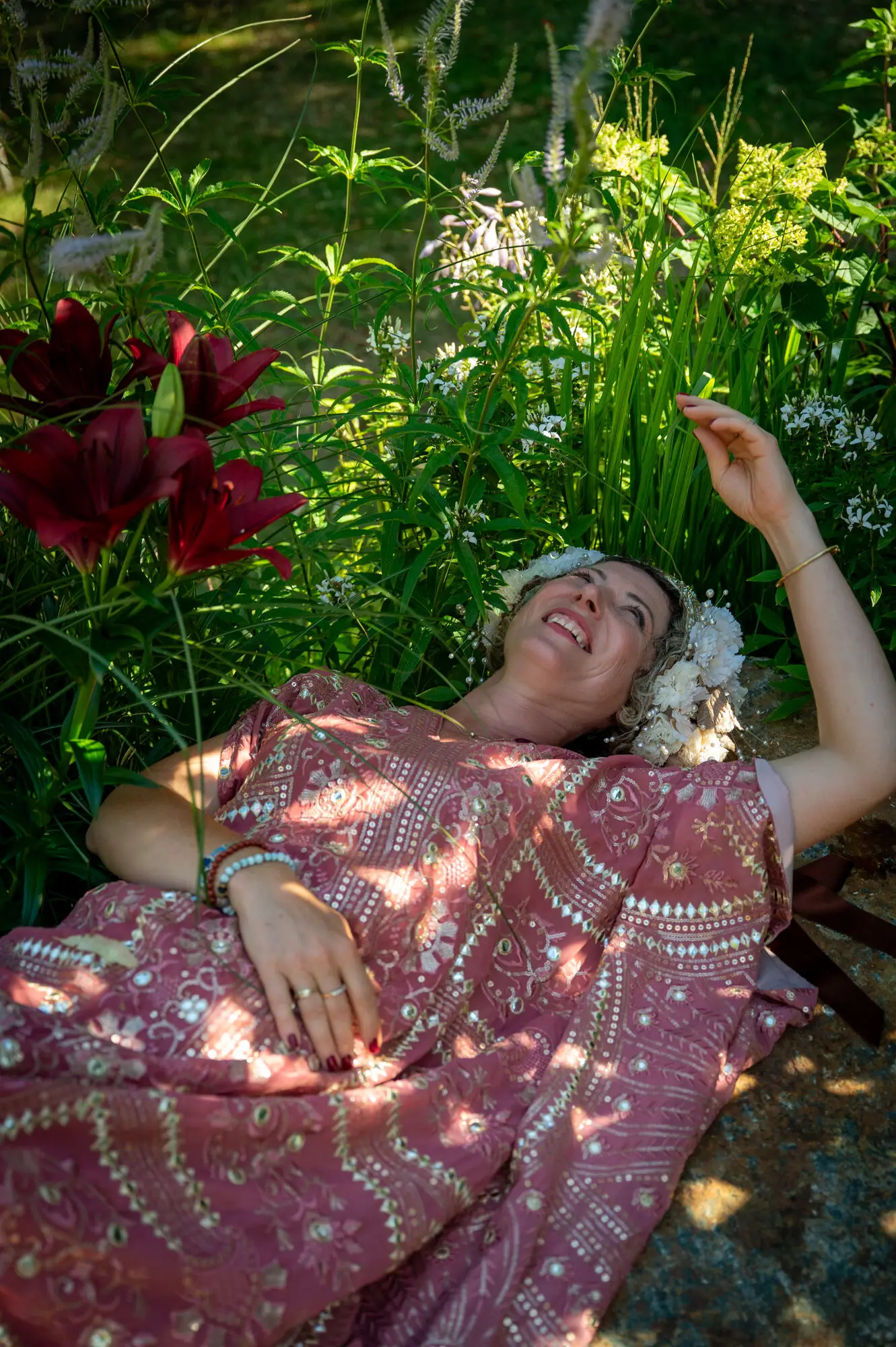 Kitti Kovacs lying back among red lilies and perennials, laughing, in a pink embroidered dress