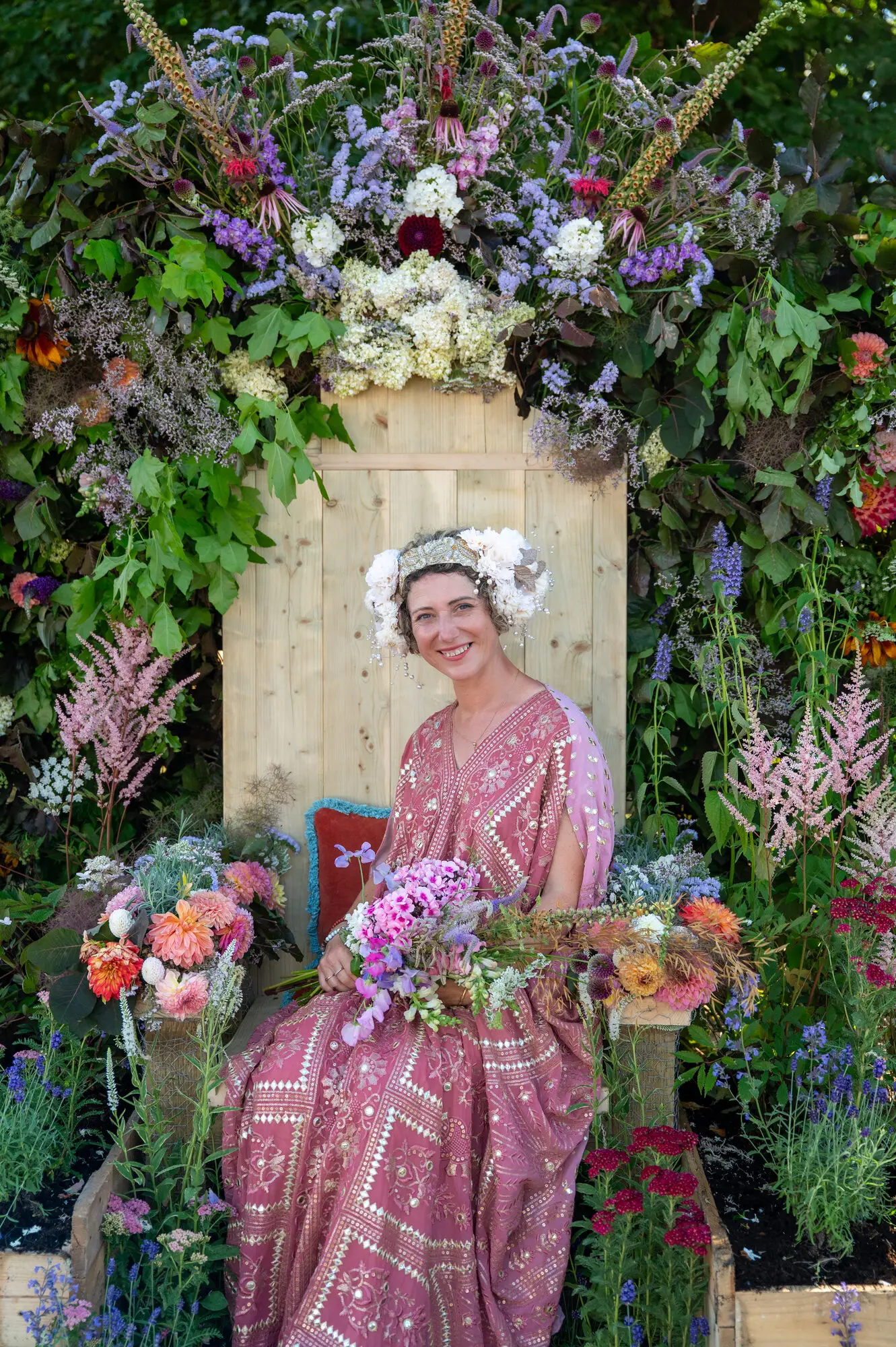 Kitti Kovacs in a flower crown and a pink embroidered dress, seated on a timber throne framed by an arch of cut flowers