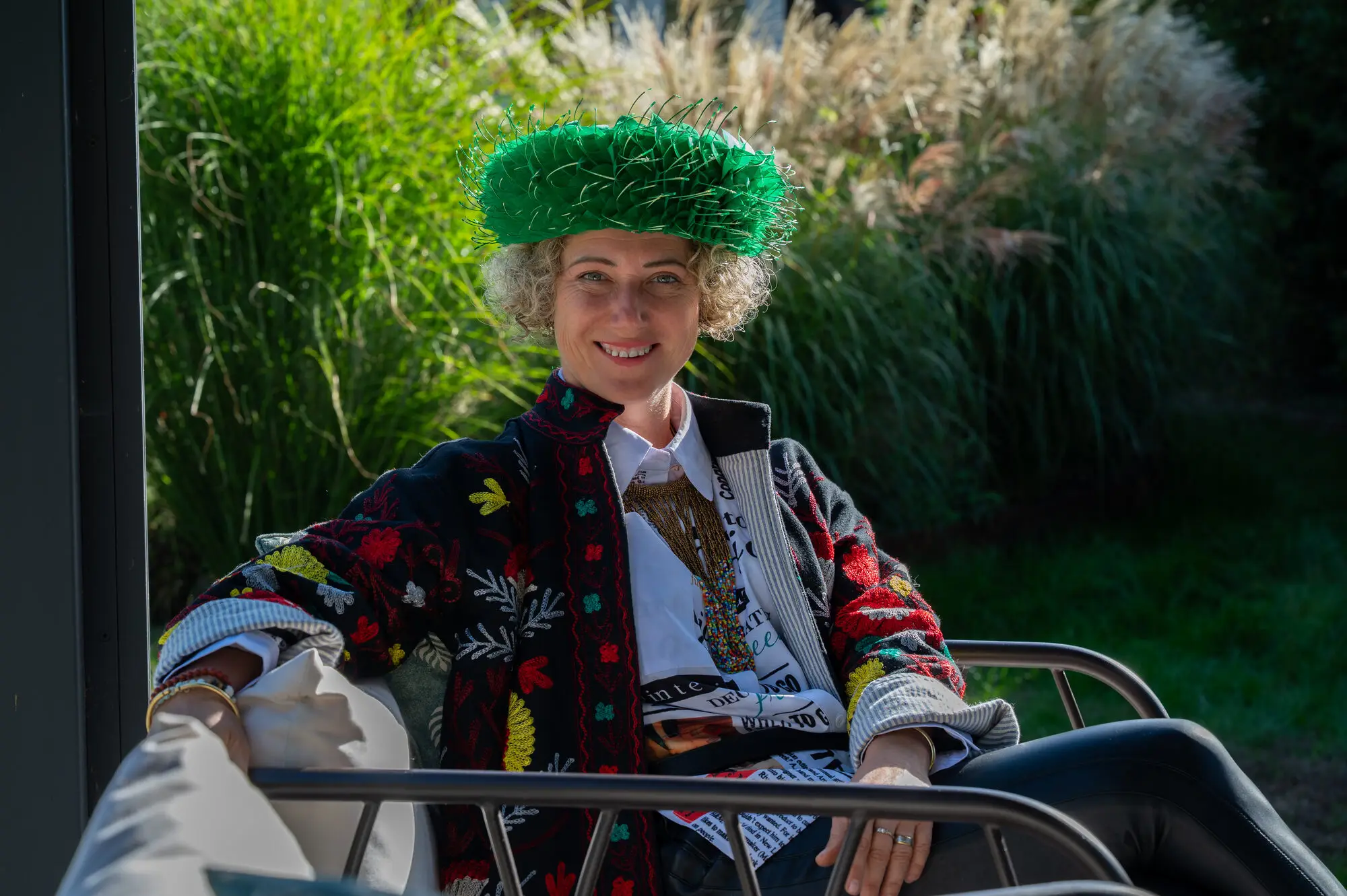 Kitti Kovacs in a green hat and embroidered jacket, sitting under a pergola with tall grasses behind her