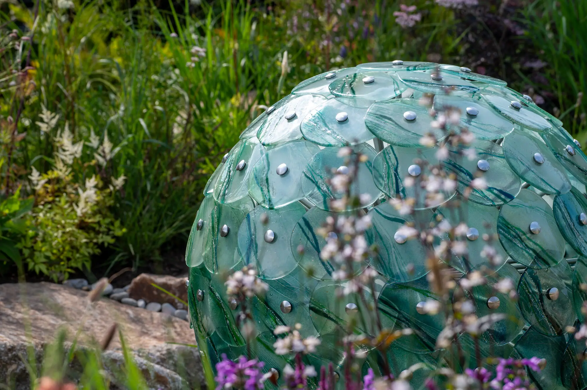 Sculptural glass water feature made of overlapping glass leaves, standing among grasses and flowering perennials