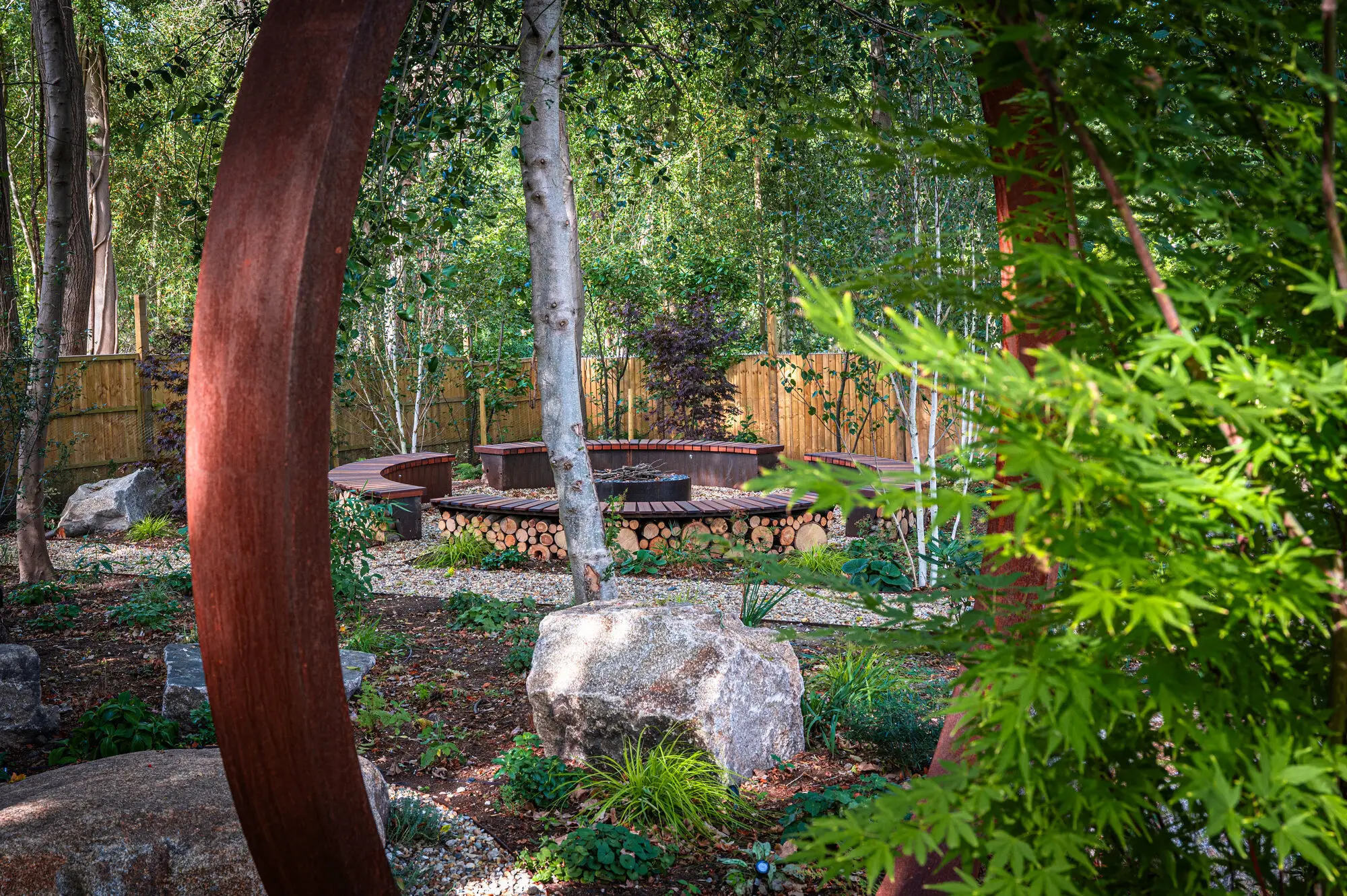 The woodland wellbeing garden seen through the corten steel moon gate, with birches, boulders and the sunken fire pit