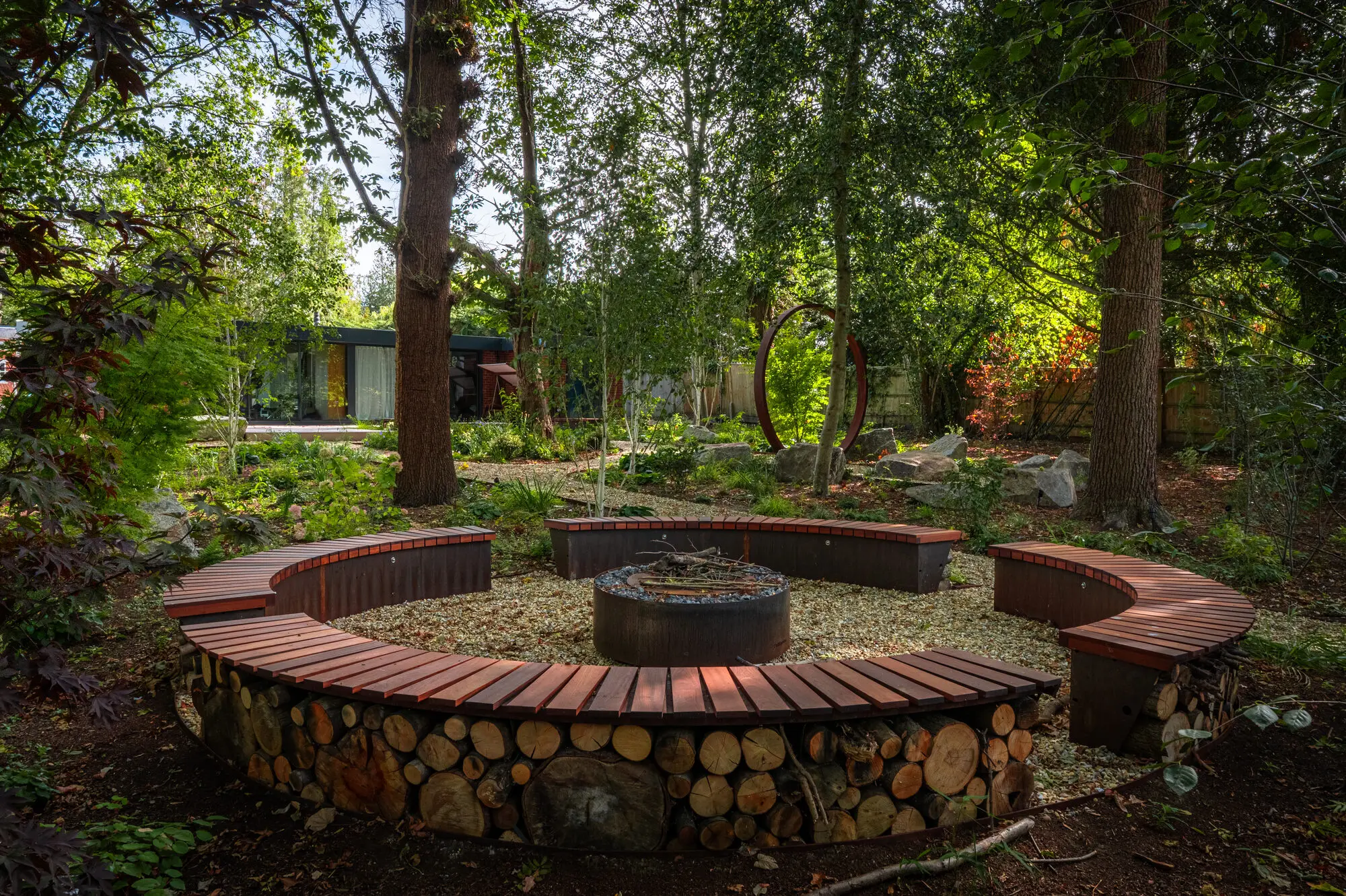 Curved timber benches over stacked logs around the fire pit, under tall trees in the woodland garden