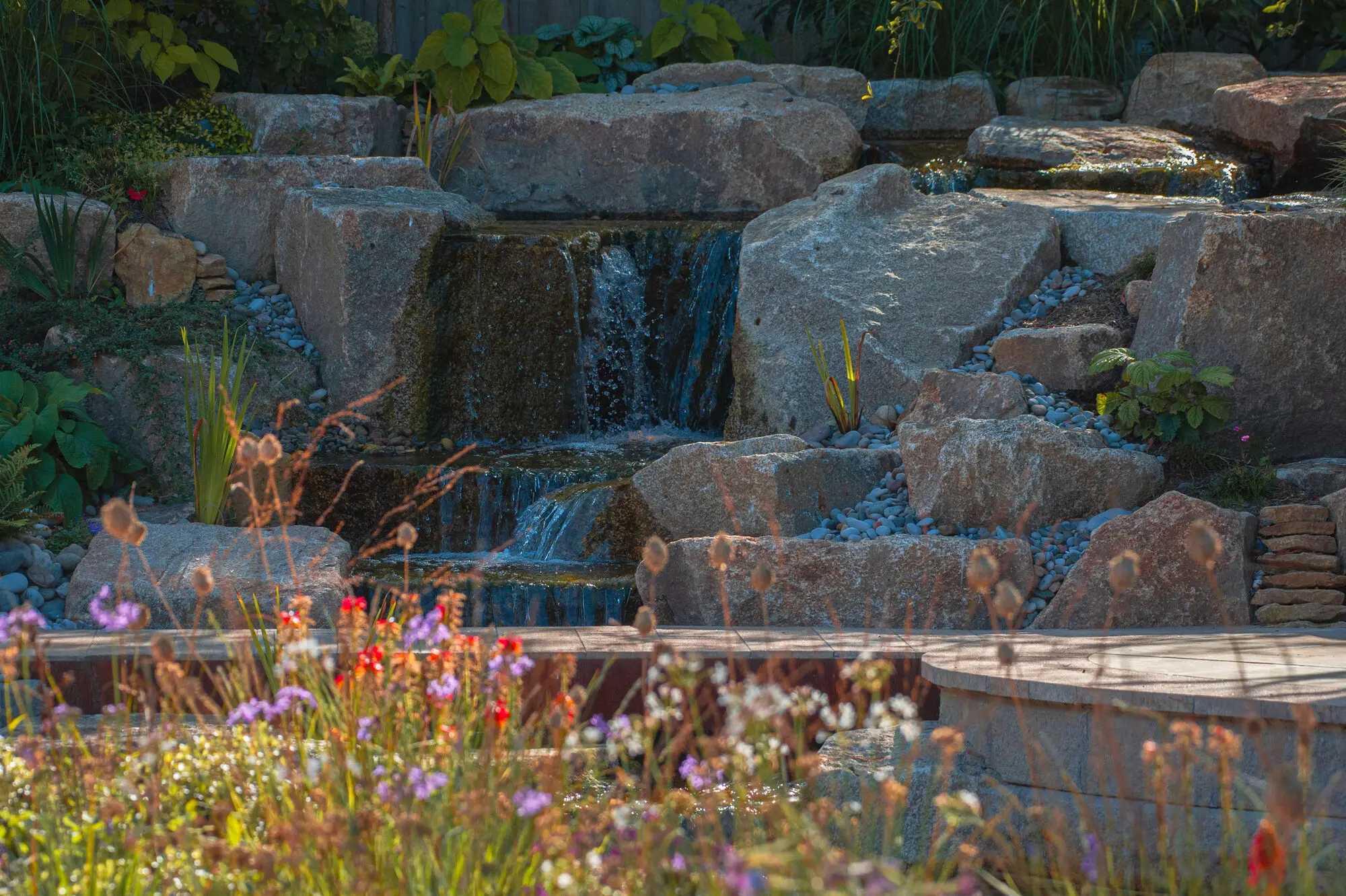 Water falling over stacked granite into the pond, behind a corten steel edge and flowering perennials