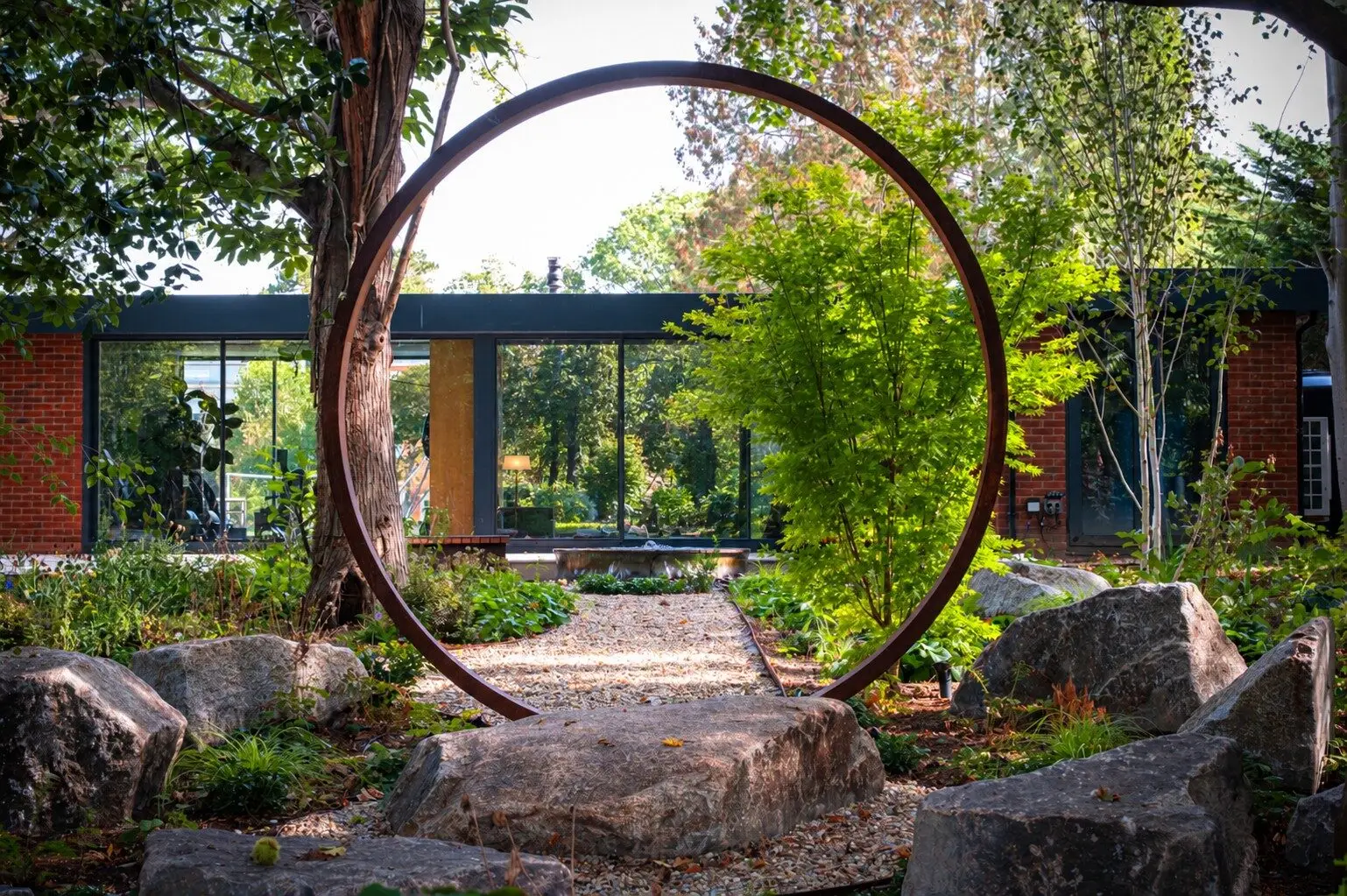 The corten steel moon gate seen head-on, framing the gravel path to the millstone water feature and the glass pool house