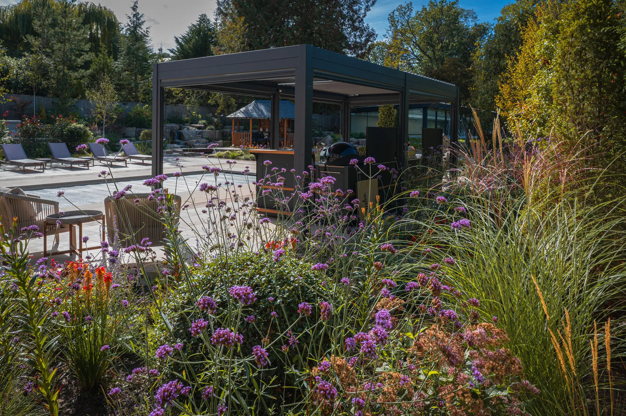 The pergola and outdoor kitchen seen through verbena and grasses, with the pool, the loungers and the Breeze House behind