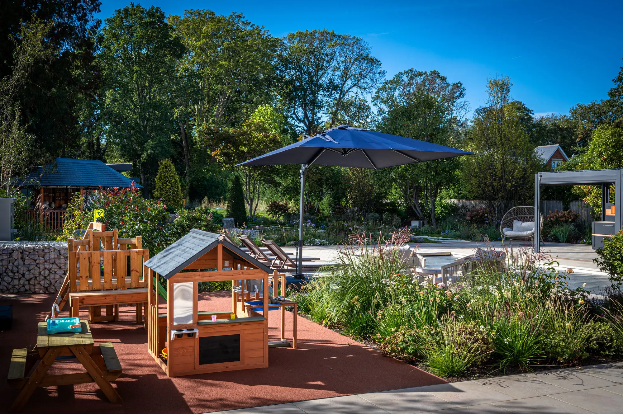 A play area with a wooden play kitchen and table on a soft surface beside the pool terrace, with the planting and the Breeze House behind