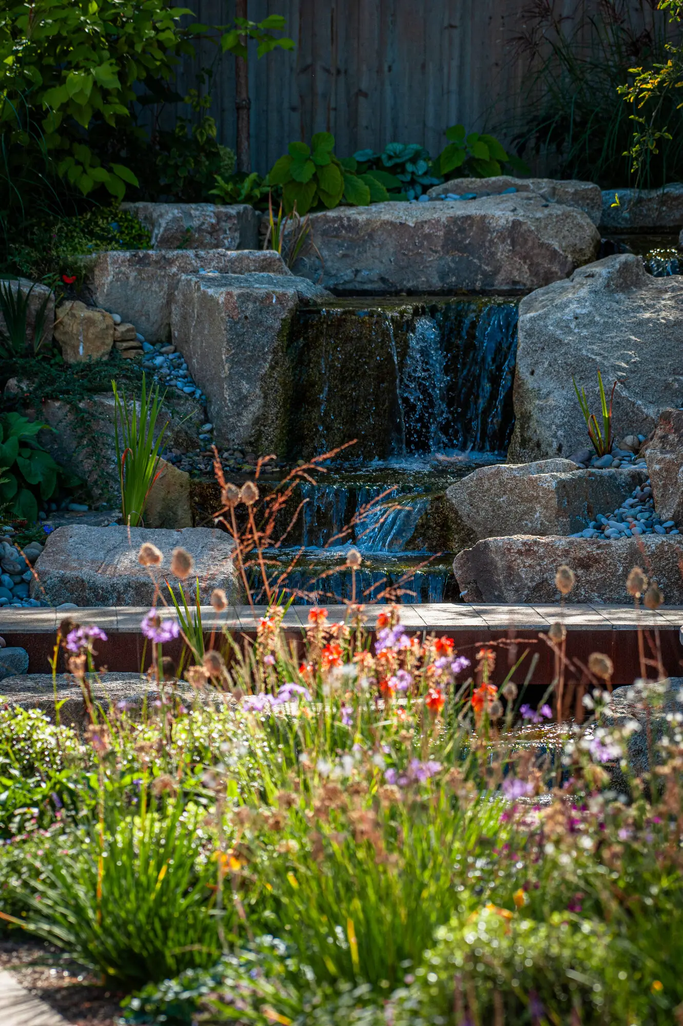 Water falling in three steps over granite boulders, with red hot pokers and alliums in front