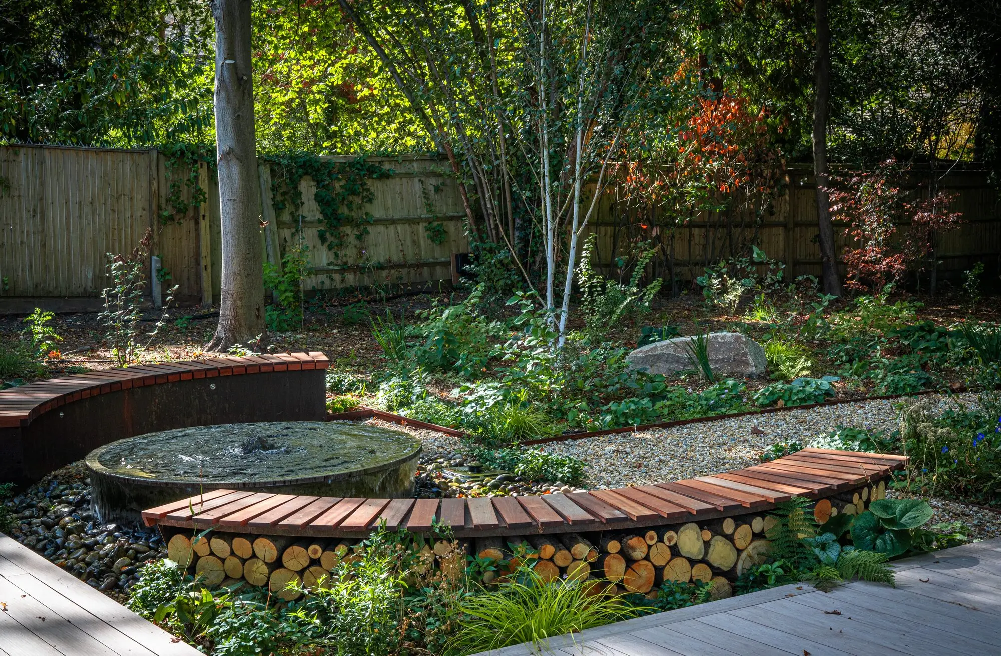 Curved benches over stacks of logs from the garden's own felled timber, around a millstone water feature in the woodland garden